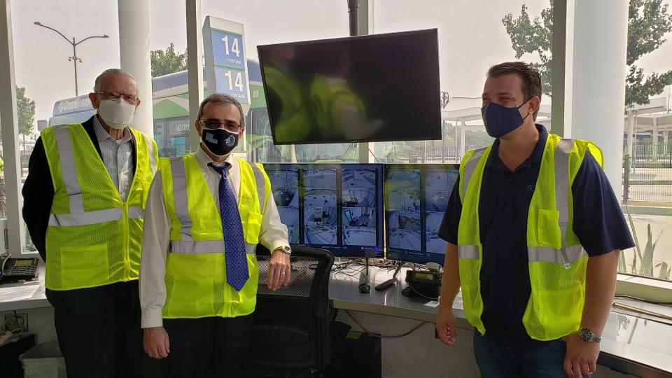 Three people in yellow safety vests stand in front of three computer screens showing video feeds.
