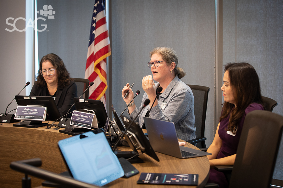 Three women at a conference table with laptops and an American flag in the background.