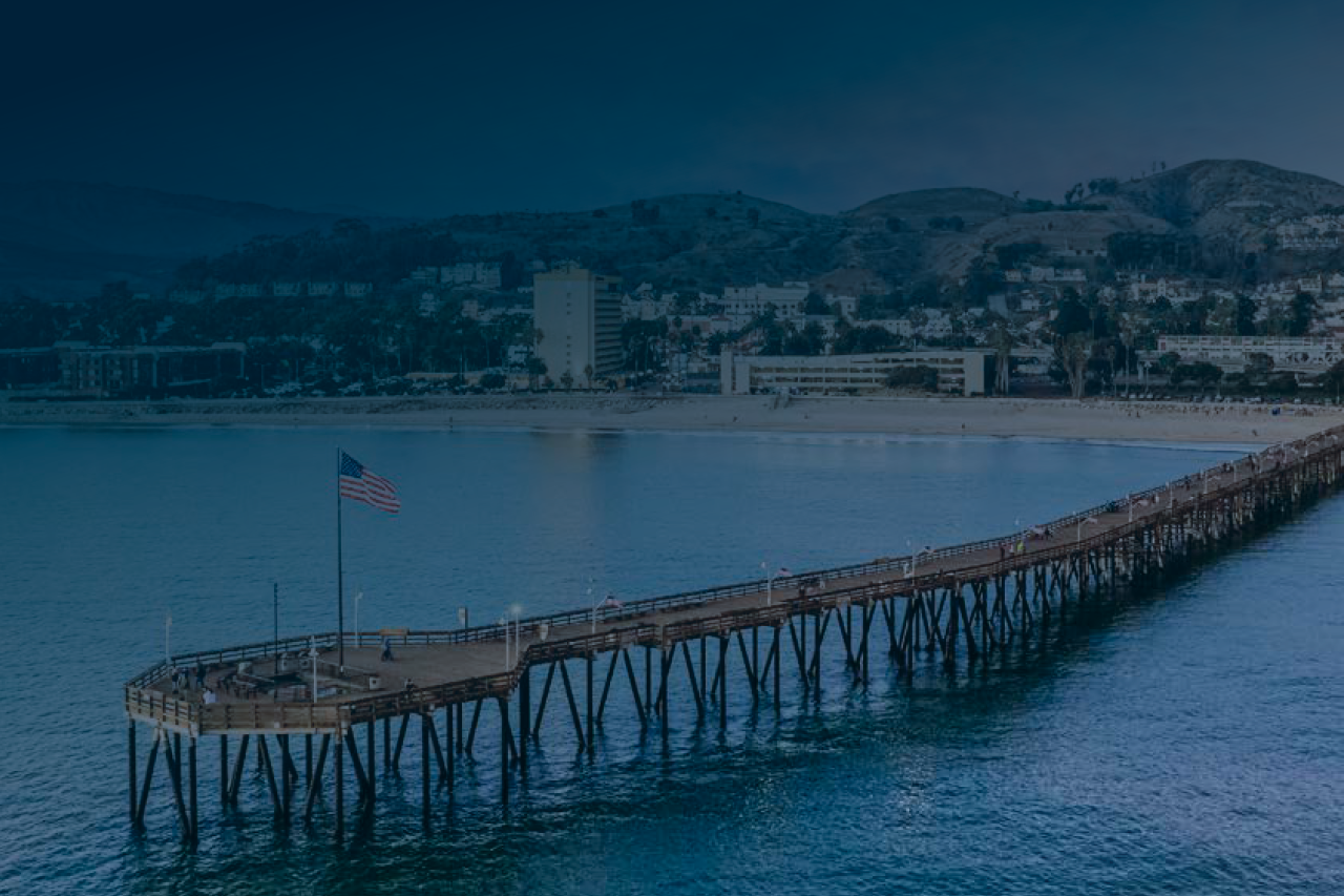Wooden pier extending into calm blue water, with distant hills under a cloudy sky.