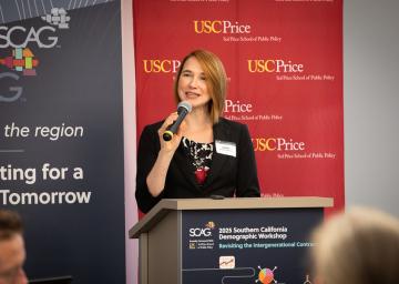 Woman speaking at a podium, red and blue banners in background.