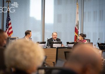 Business meeting with three people at a conference table, flags in background.