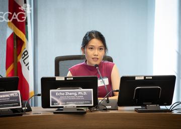 Woman speaking at a conference with multiple monitors on the table.