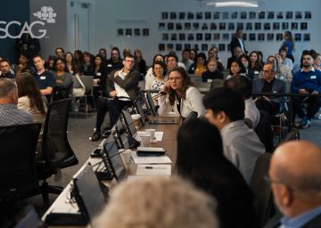 Meeting with diverse people seated at tables, discussing attentively in a brightly lit room.