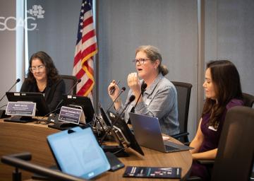 Three women at a conference table with laptops and an American flag in the background.