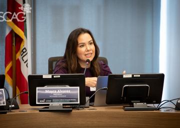 Woman speaking at a conference table with monitors and flags behind.