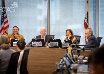 Four people seated at a conference table, engaged in discussion. U.S. and state flags behind them.