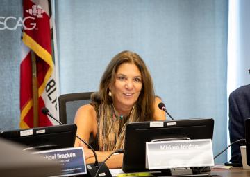 Woman speaking at a conference table, flag in background.