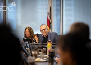 Man and woman at a conference table, smiling, with laptops and a flag behind.