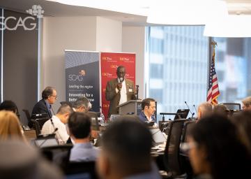 Man speaking at conference with seated audience, flags, and banners.