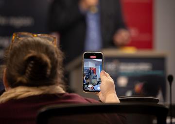 Audience member films speaker on phone in conference setting.