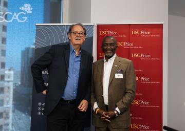Two men in suits smiling in front of SCAG and USC Price banners.