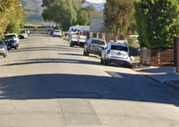 Street lined with parked cars and trees, blue recycling bin by the curb.