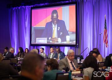 Conference with attendees seated, large screen displays a speaker, U.S. flag in background.