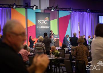 Conference audience facing colorful stage with speaker, some standing.