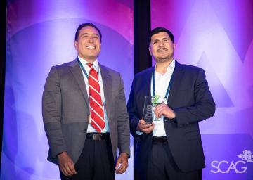 Two men in suits, one holding an award, stand against a blue and purple backdrop.