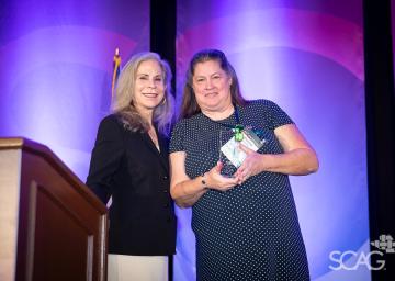 Two women stand together, one holding a glass award, against a purple backdrop.