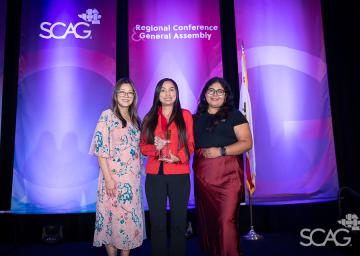 Three women smiling on stage with purple conference banners in the background.