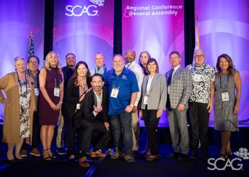 Group of people smiling on stage at a conference with purple backdrop.