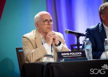 Seated man in a beige suit at a conference, appears thoughtful.