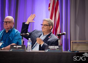 Two men seated at a panel, one raising his hand, with a U.S. flag behind them.