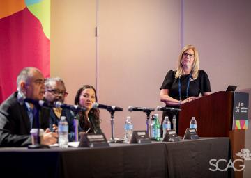 Panel discussion with four seated speakers and a woman standing at a podium.