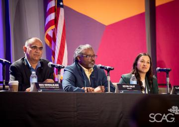Panel of three speakers seated with microphones in front of a colorful backdrop and a flag.