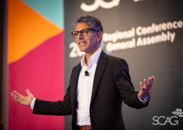 Man speaking energetically on stage at a conference, colorful backdrop.