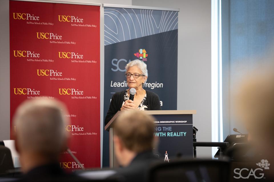 A person standing at a podium in front of a blue SCAG banner and a red USC banner addresses the attendees at the 2024 Southern California Demographic Workshop.